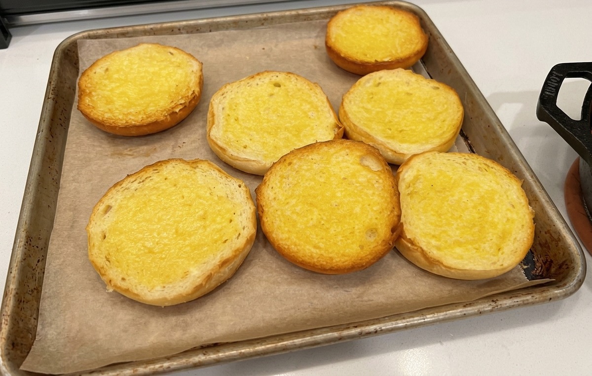 Metà di panini per hamburger imburrate e disposte su carta forno in una teglia.