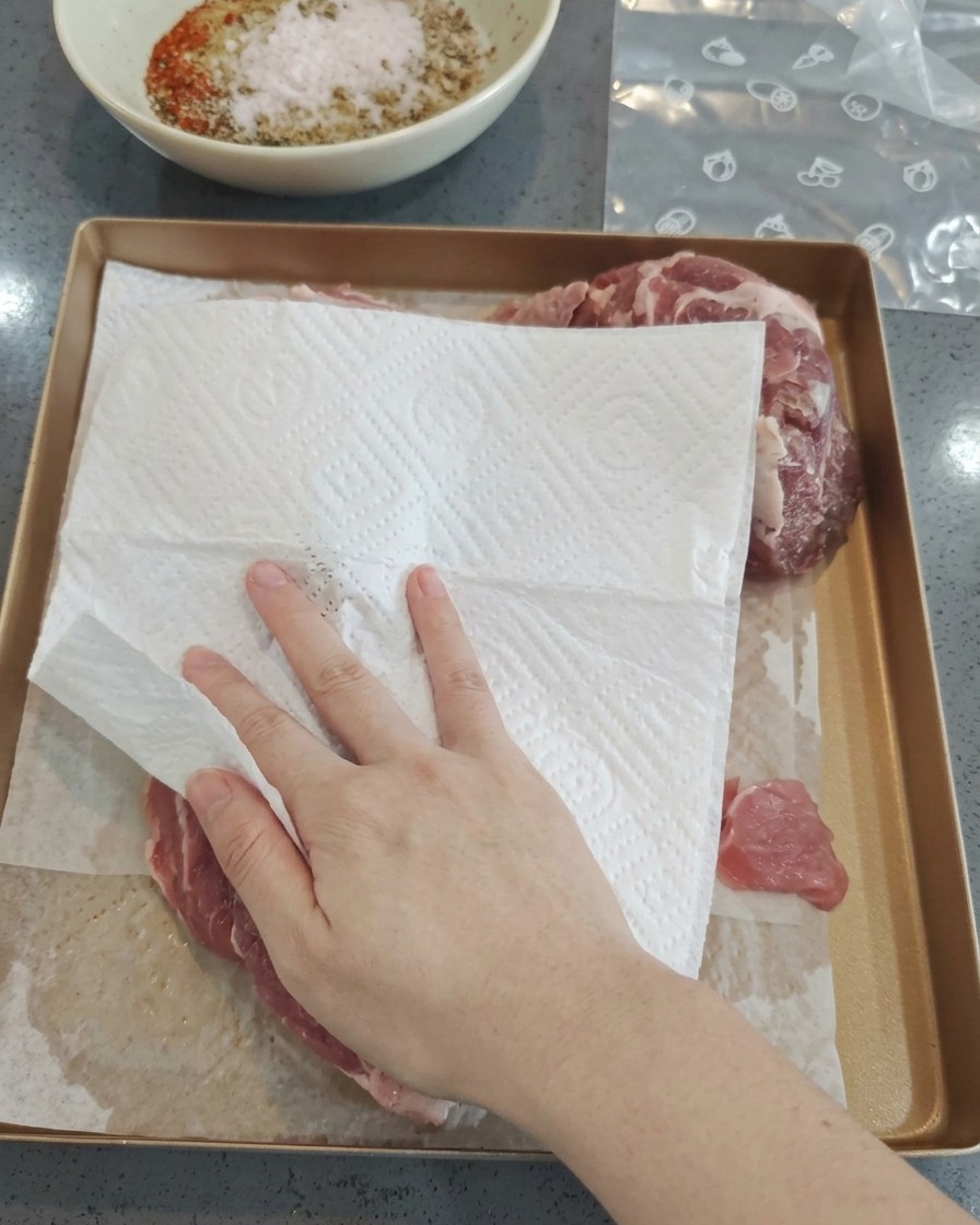 Una mano usa carta assorbente per tamponare pezzi di maiale crudo su una teglia dorata.