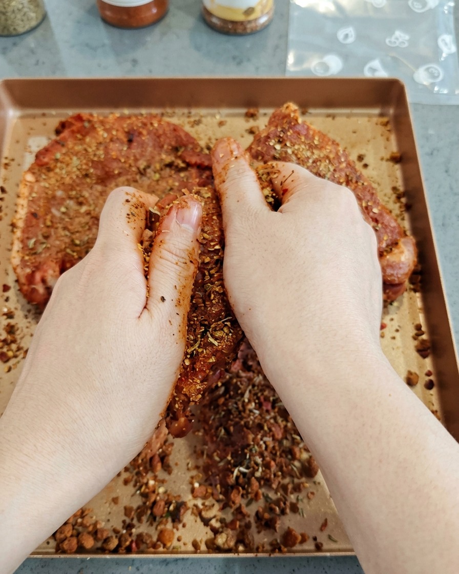 Mani che massaggiano un mix di spezie scuro e saporito su grandi pezzi di maiale crudo su una teglia.
