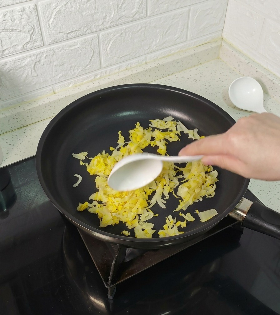 Una mano con un cucchiaio bianco mescola i crauti gialli in una padella antiaderente nera.