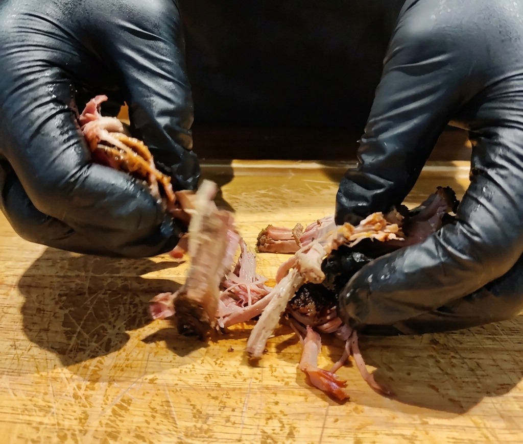 Mani guantate che sfilacciano un pezzo di coppa di maiale arrosto su un tagliere di legno.