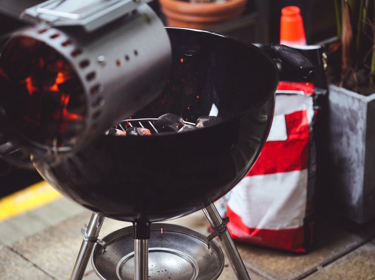 Carbone ardente e rosso che viene versato da una ciminiera di metallo sul fondo di un barbecue nero.