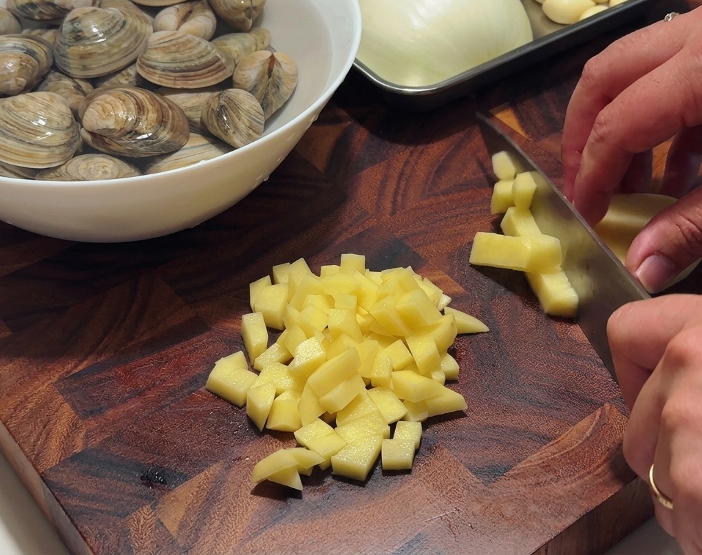 Mani che usano un coltello da chef per tagliare patate sbucciate a cubetti su un tagliere di legno scuro.