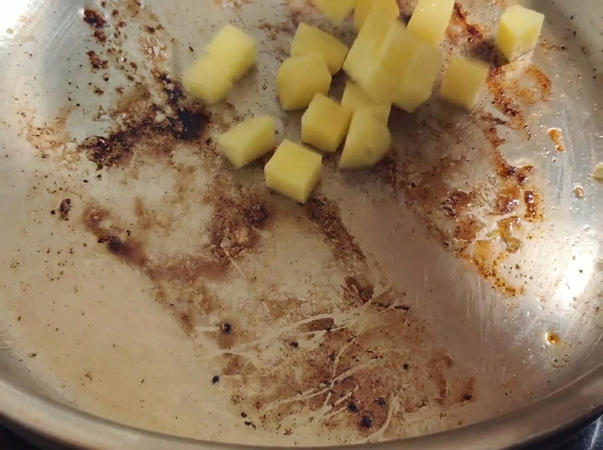 Cubetti di patate gialle che cadono in una padella calda con il grasso del bacon e piccoli residui dorati.