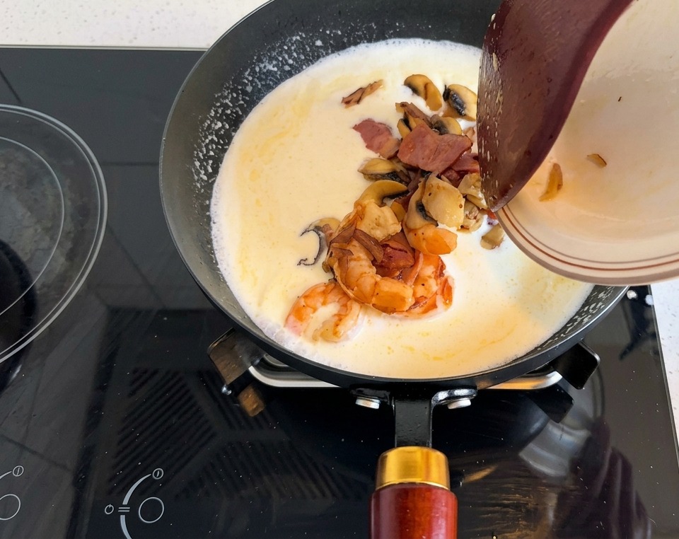 Un piatto con gamberi, pancetta e funghi versato nella base cremosa in padella.