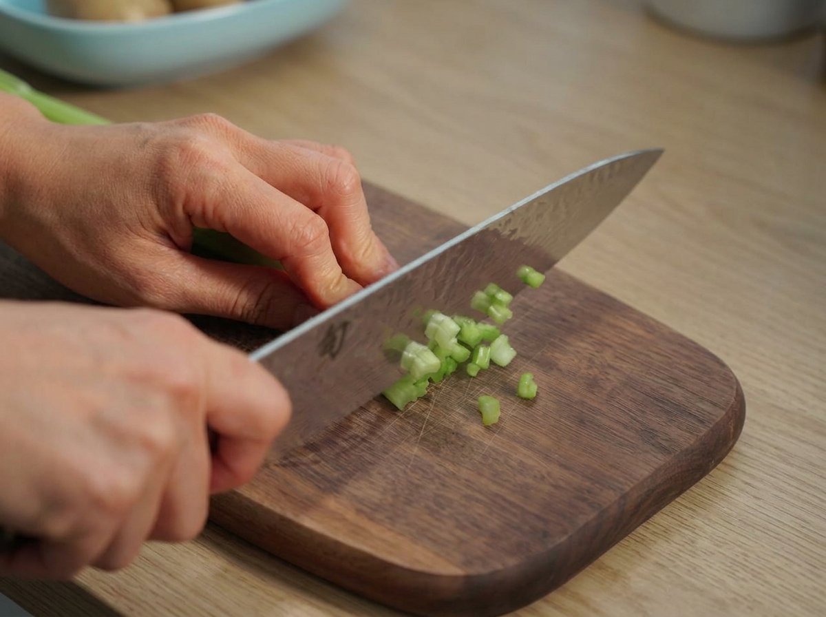 Mani che usano un coltello da chef per tagliare il sedano su un tagliere di legno.