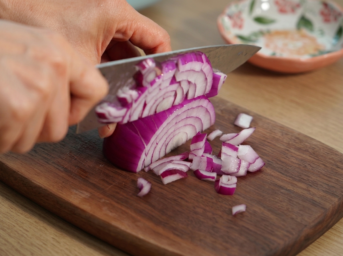 Mani che usano un coltello da chef per tritare finemente una cipolla rossa su un tagliere.