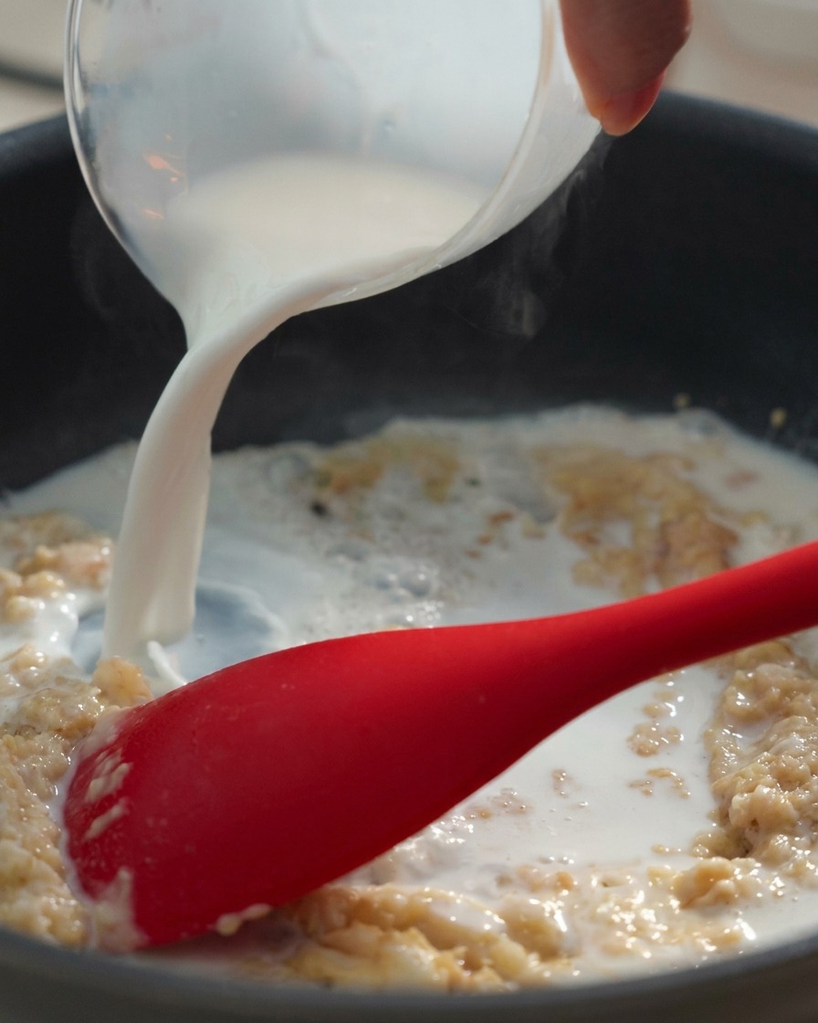 Latte versato in una padella con un composto denso di burro e farina, mescolato con una spatola rossa.