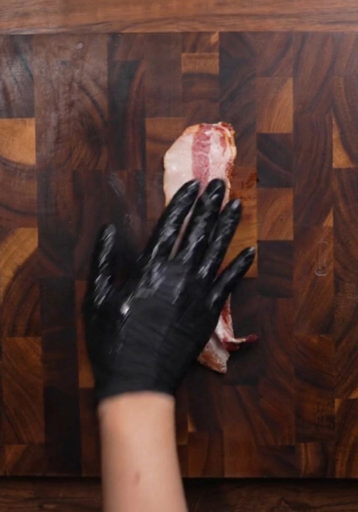 Una mano con guanto su una fetta di bacon crudo mentre si prepara a affettarlo su un tagliere.