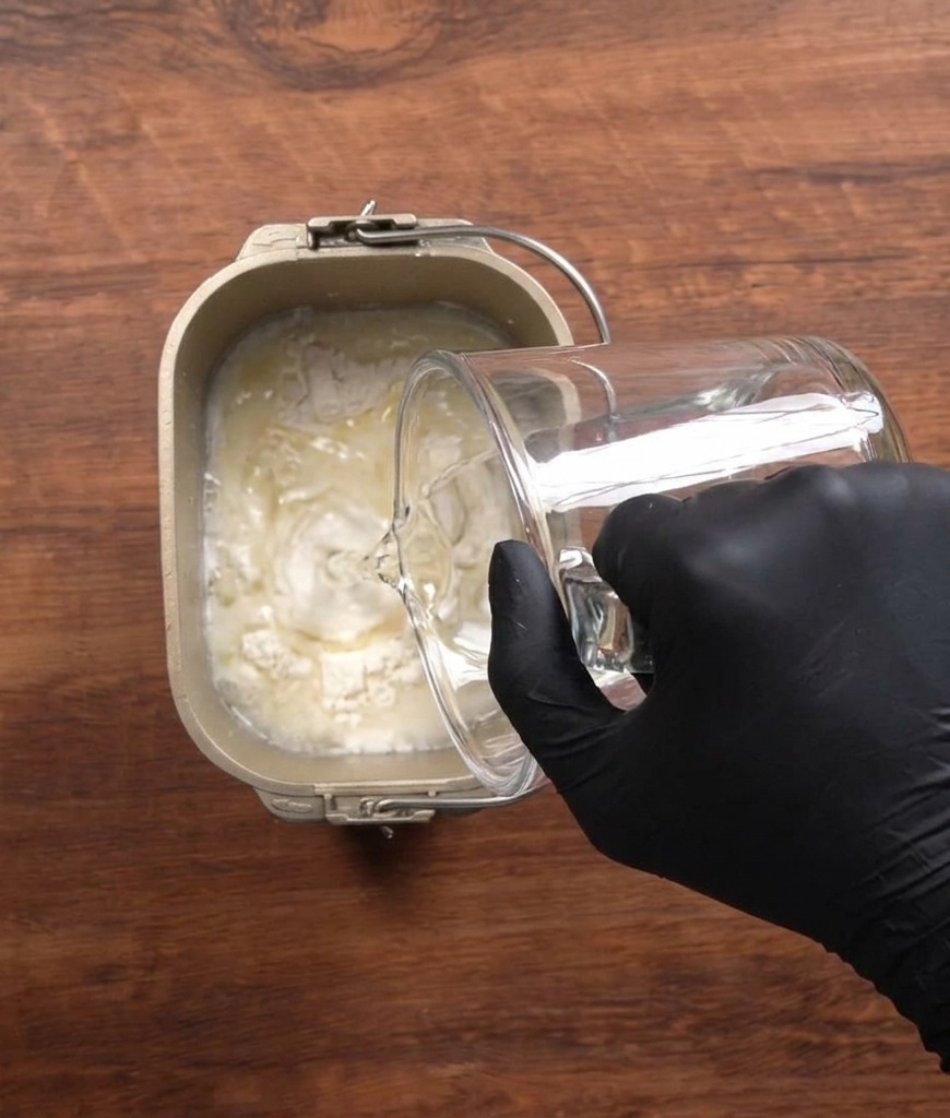 Acqua versata da un misurino di vetro nel cestello di una macchina del pane con farina e altri ingredienti.