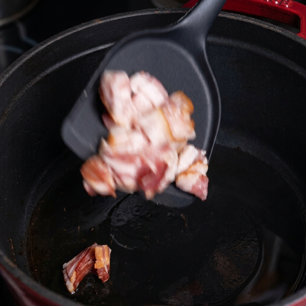 Pezzetti di bacon crudo che cadono da una spatola nera in una pentola di ghisa calda.