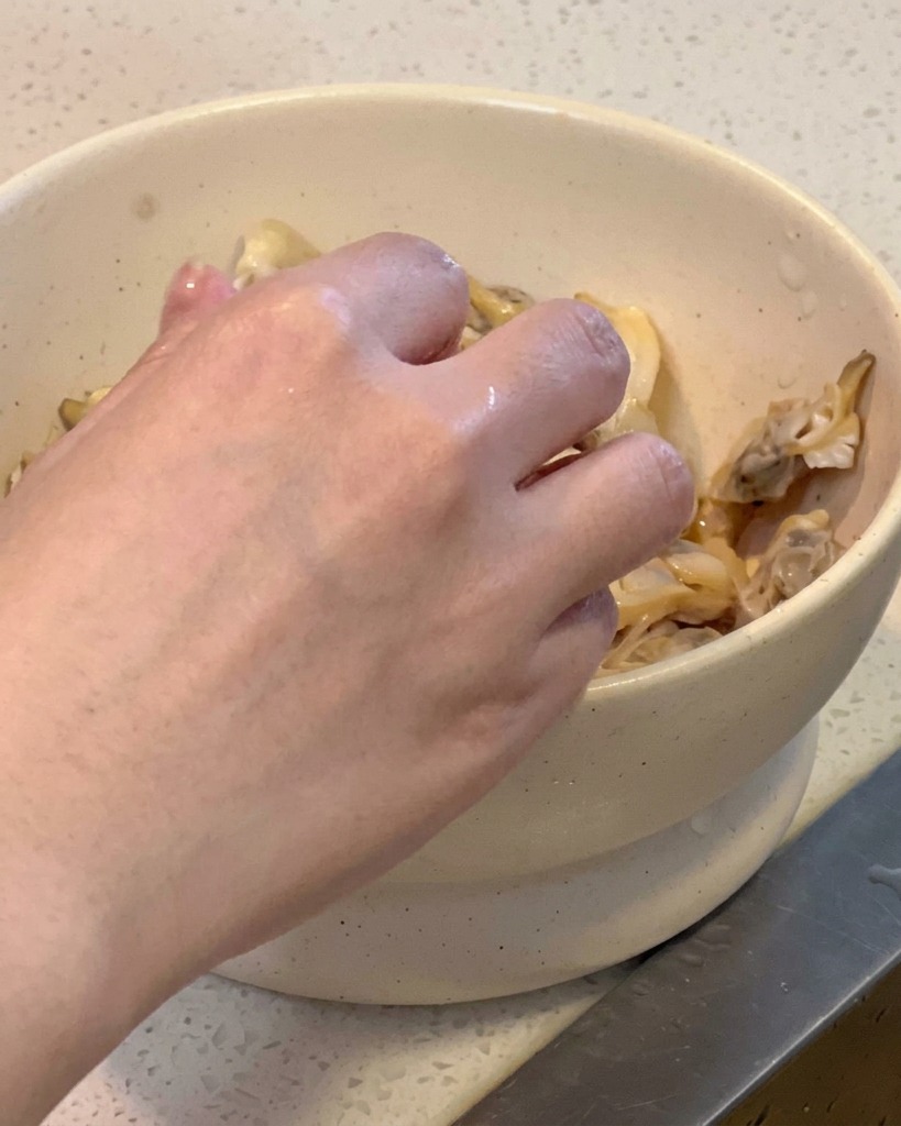 Una mano che rimuove la carne della vongola dal guscio mettendola in una ciotola bianca.