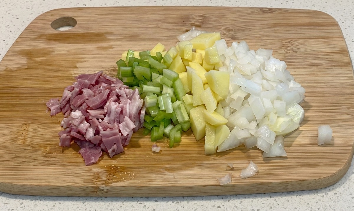 Dadini di pancetta, sedano verde, patate gialle e cipolle bianche disposti ordinatamente su un tagliere di legno.