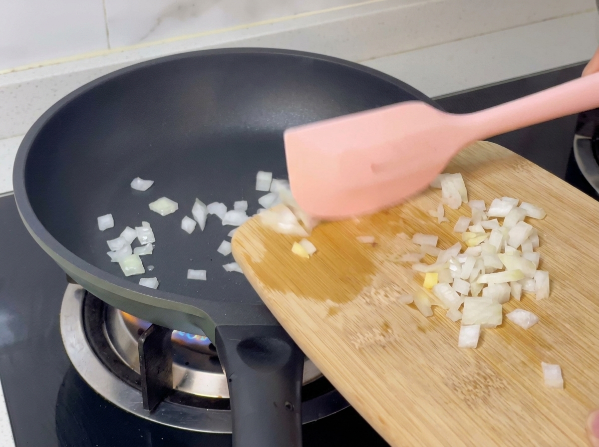 Dadini di cipolla bianca versati da un tagliere in una padella calda con una spatola rosa.