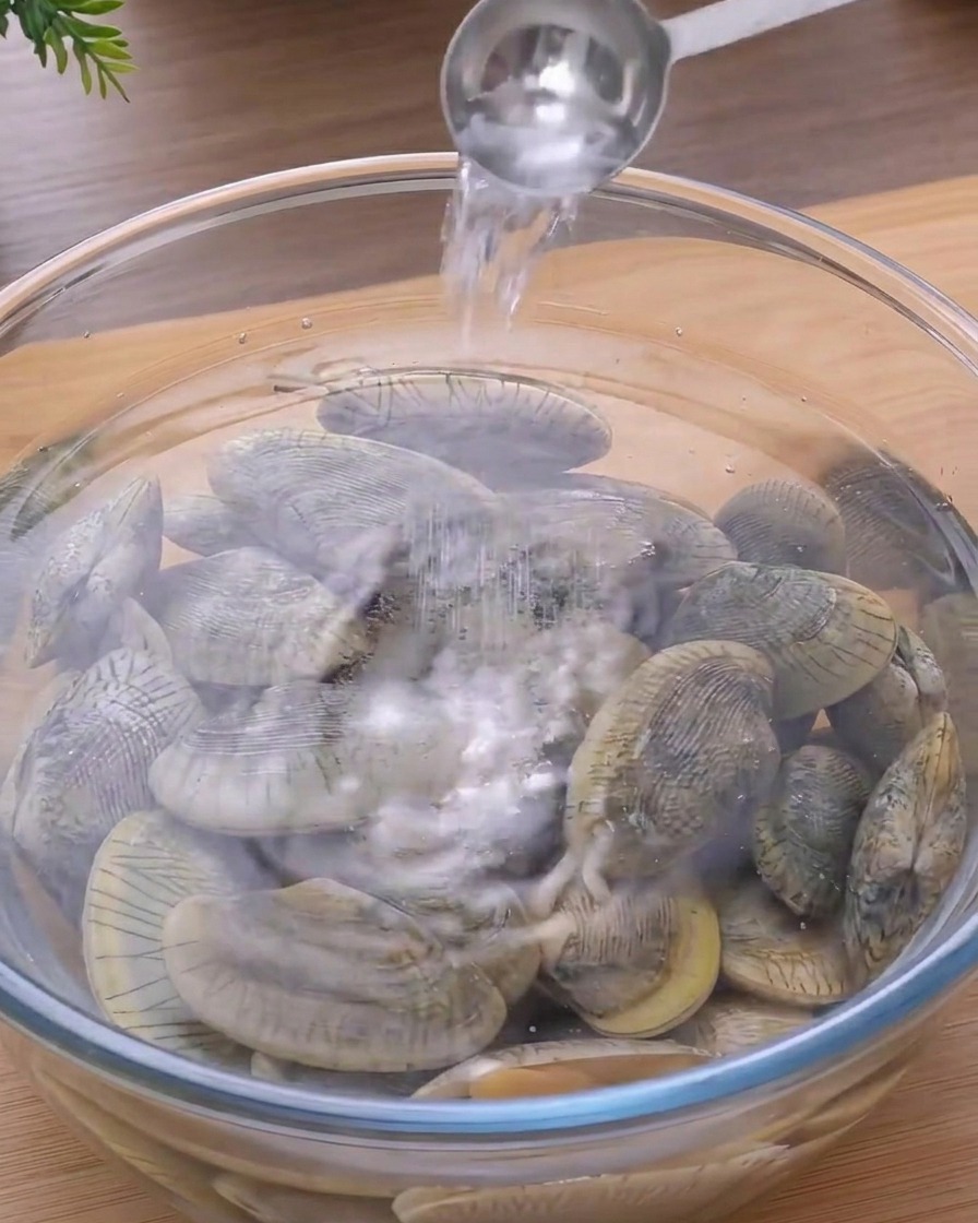 Un cucchiaio di metallo che versa sale in una ciotola di vetro con acqua e vongole fresche.