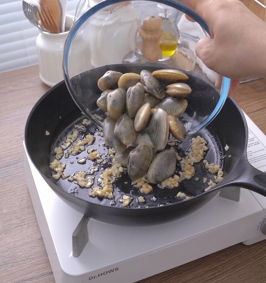Vongole versate da una ciotola di vetro in una padella calda con aglio e burro.