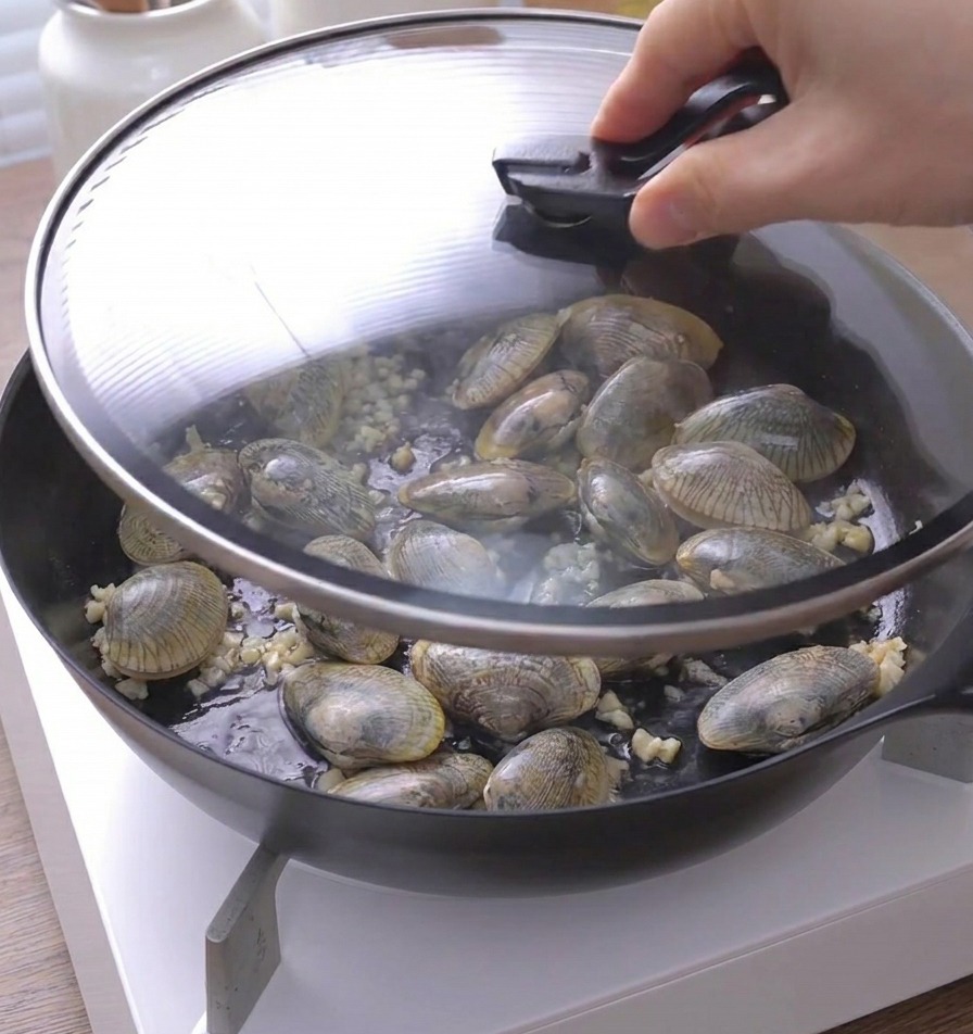 Una mano che posiziona un coperchio di vetro sopra la padella con le vongole.