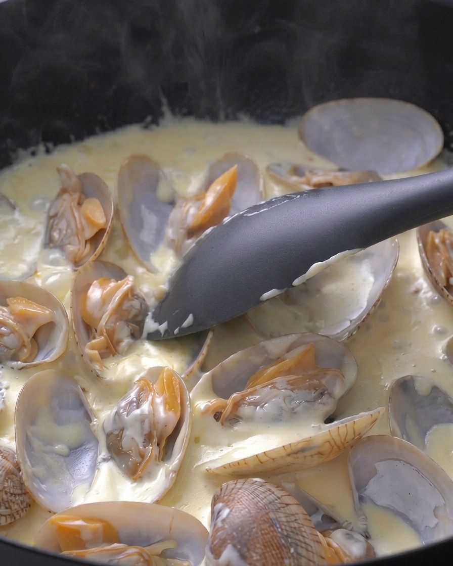 Una spatola nera che mescola delicatamente le vongole in una salsa bianca cremosa in padella.