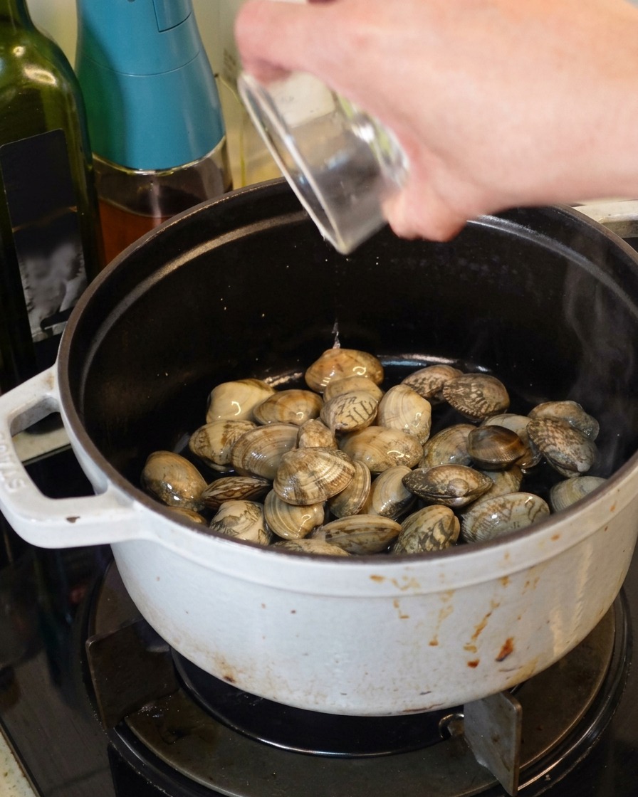 Versare del vino bianco da un bicchierino in una pentola bianca in ghisa piena di vongole fresche.