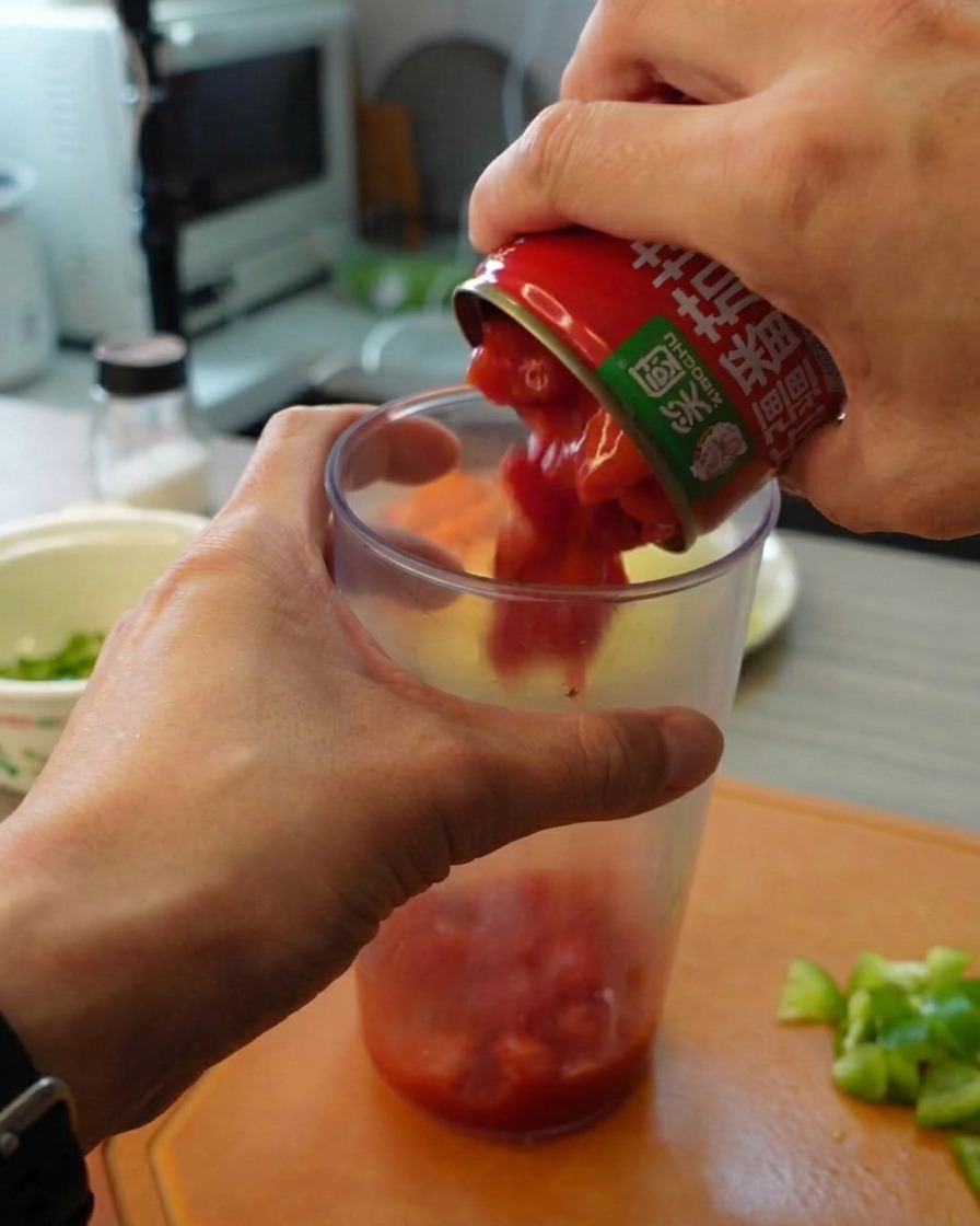 Mani che versano pomodori pelati a pezzettoni da una lattina in un alto contenitore trasparente per frullare.