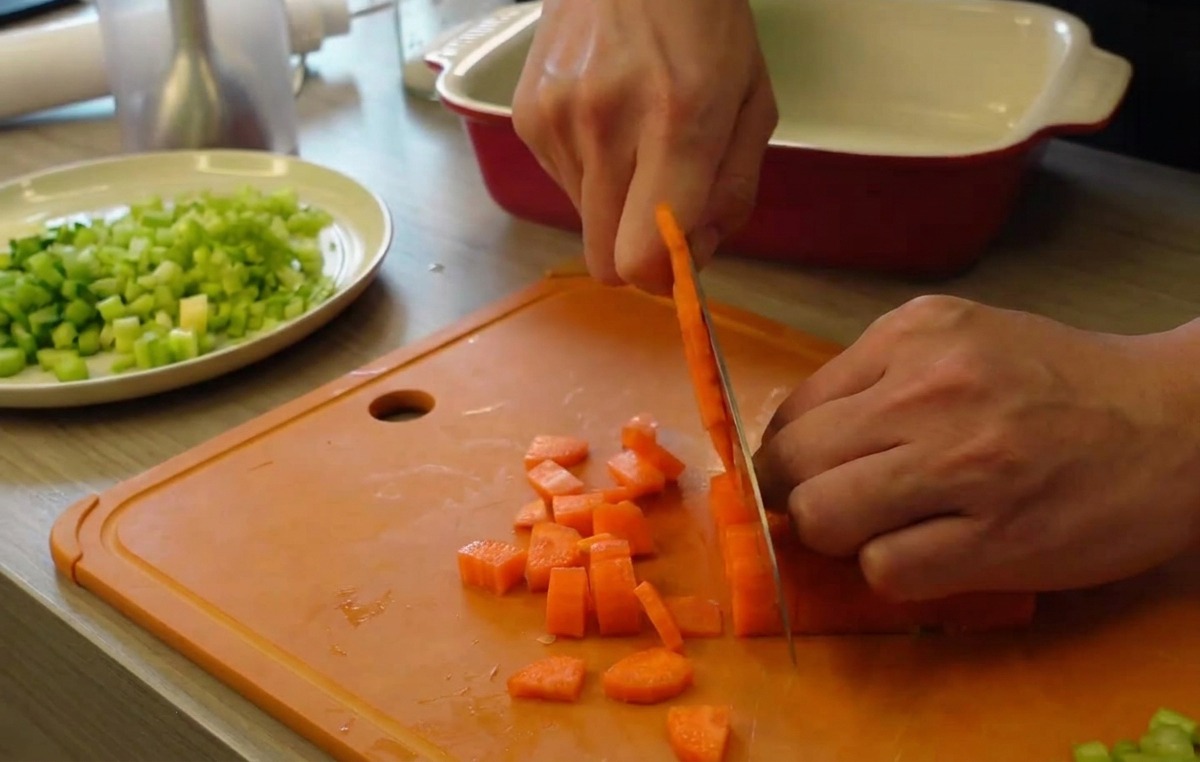Mani che usano un coltello per tagliare carote arancioni a piccoli cubetti su un tagliere arancione, con un piatto di sedano a pezzetti sullo sfondo.