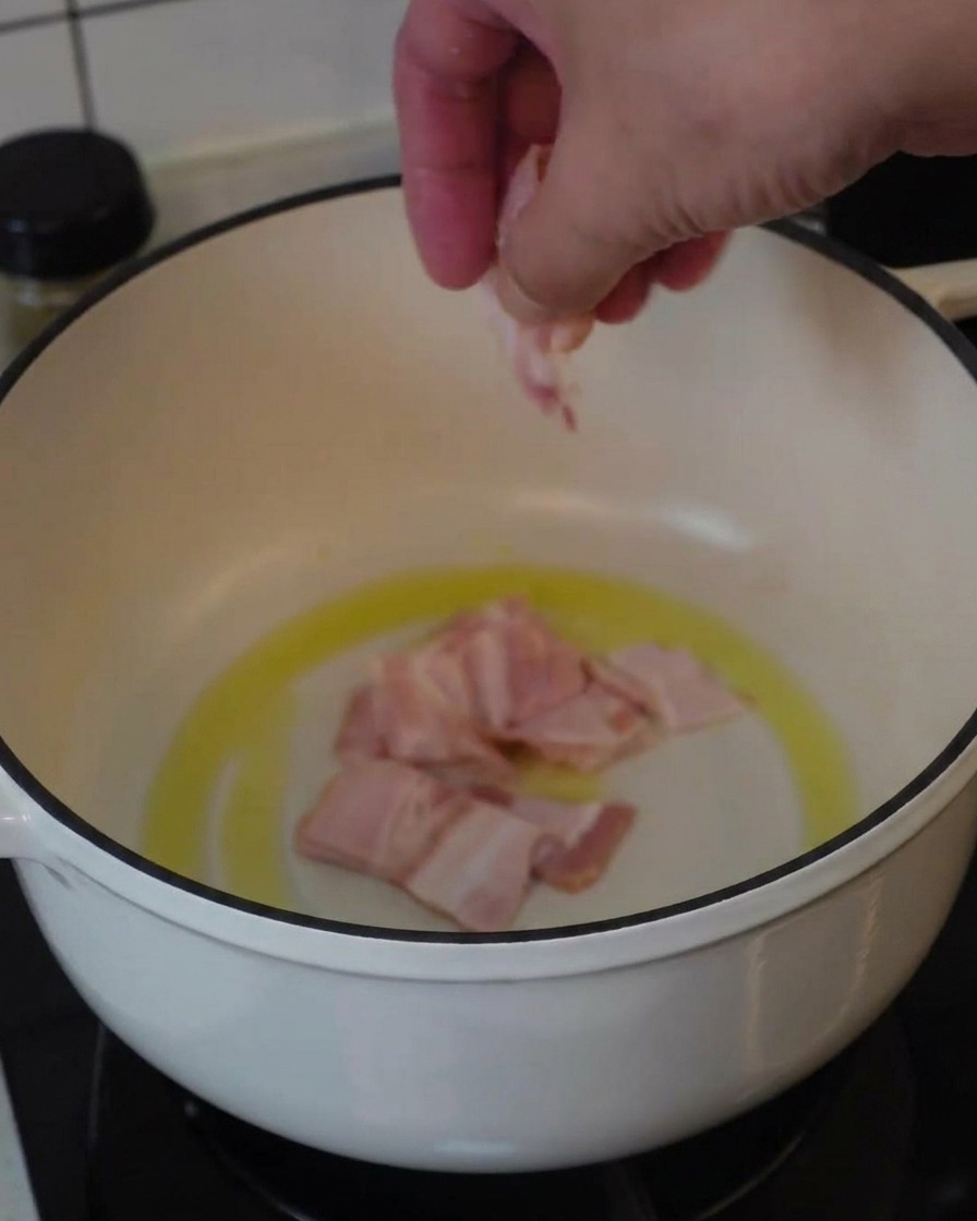Una mano che mette pezzi di bacon crudo in una pentola bianca con un po di olio per iniziare a friggerli.