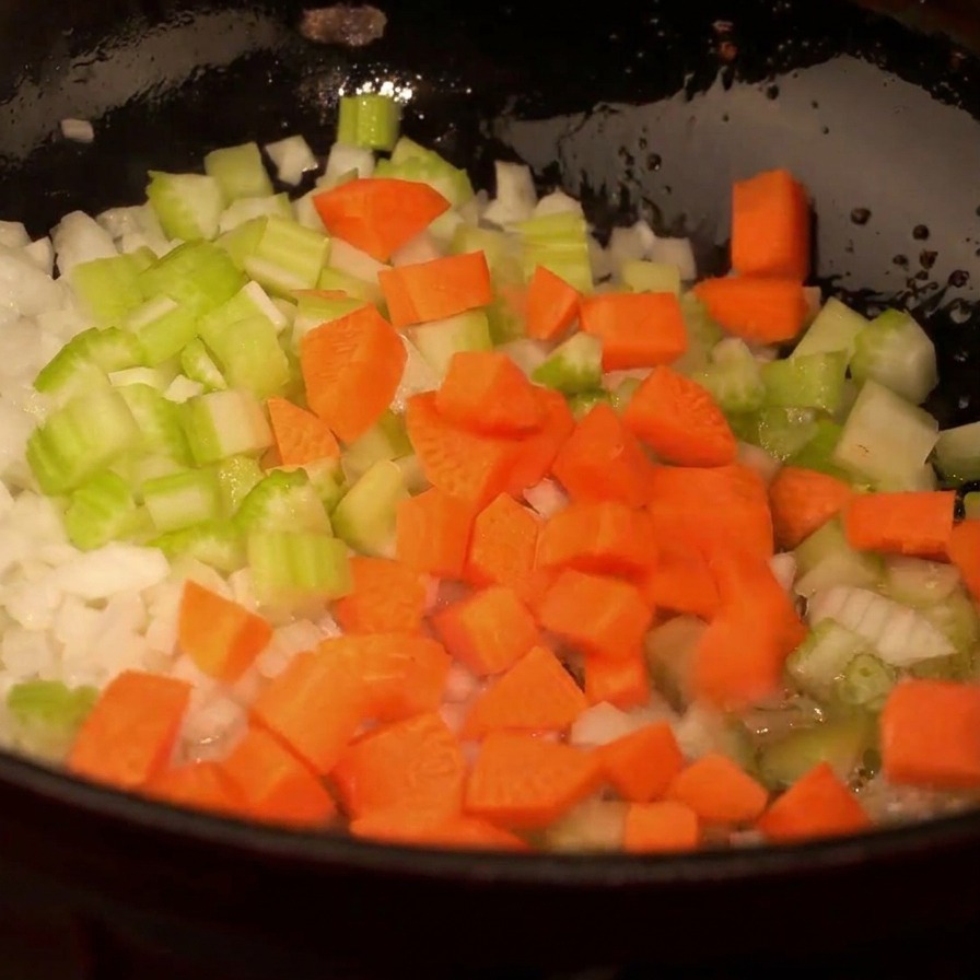Vista dallalto di una padella nera con cipolle bianche tritate, sedano verde e cubetti di carota arancione in soffritto.