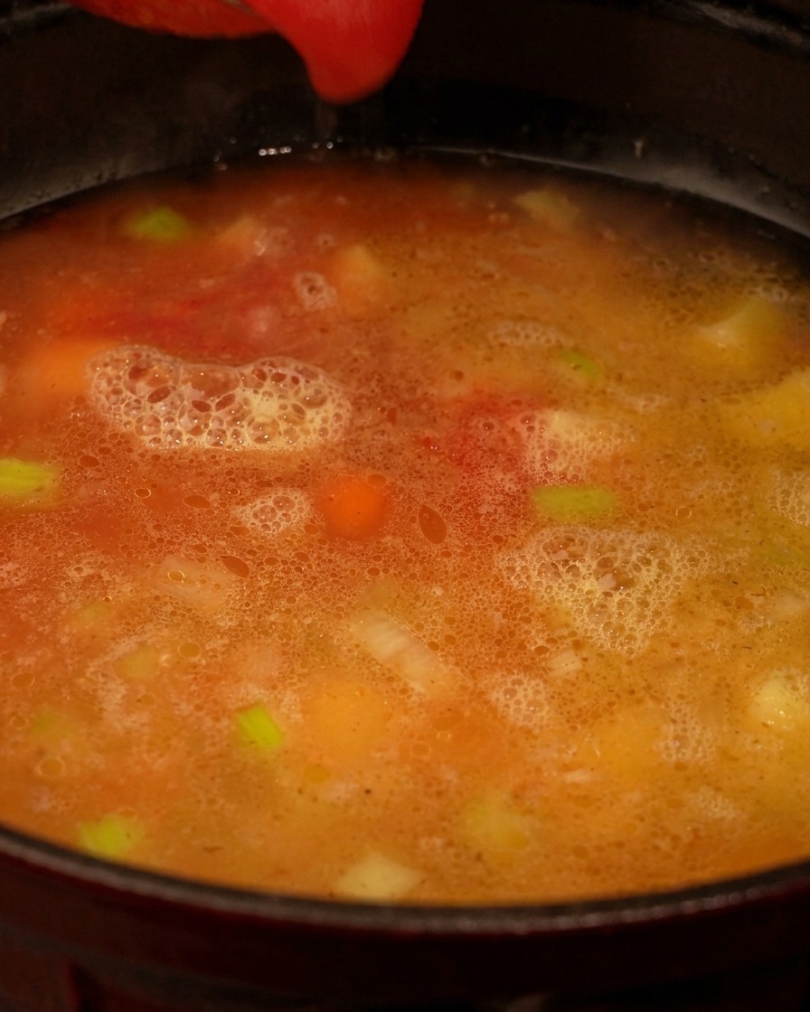 Passata di pomodoro rossa versata in una pentola contenente brodo chiaro e pezzi visibili di sedano e carote.