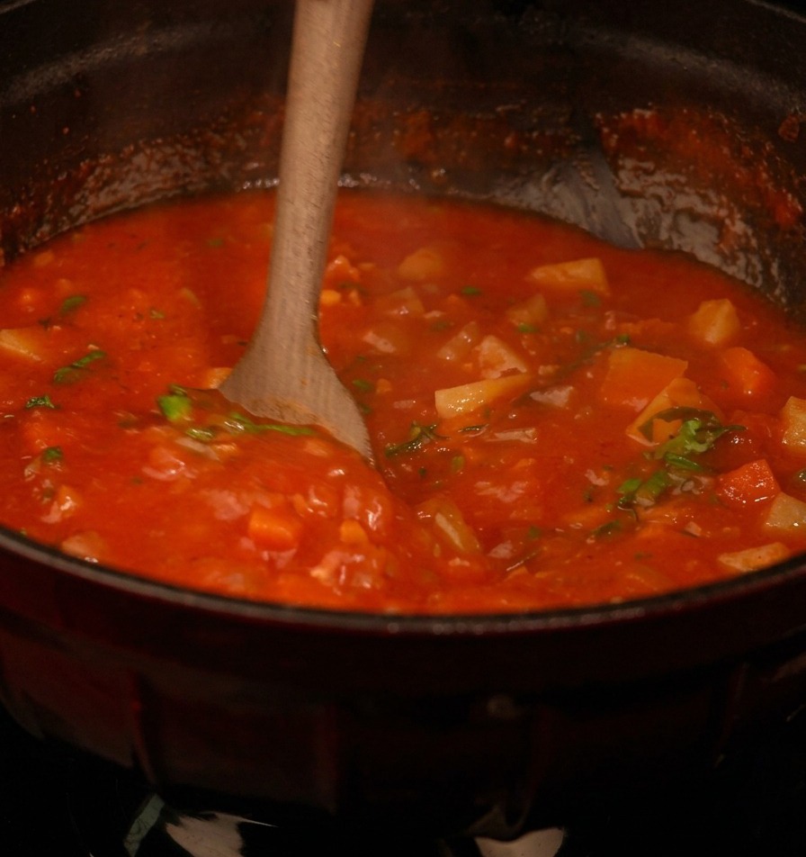 Cucchiaio di legno che mescola una densa zuppa di pomodoro piena di verdure e pezzetti di prezzemolo verde fresco.
