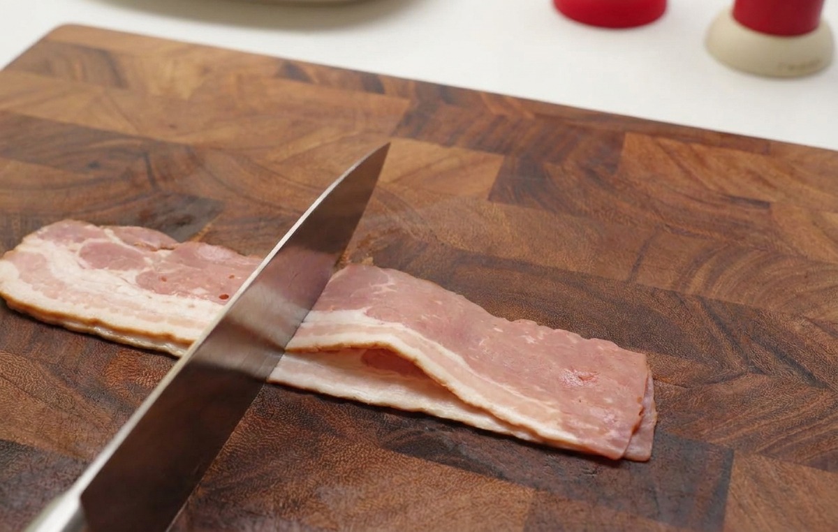 Mani che usano un coltello affilato per tagliare strisce di pancetta cruda in piccoli pezzi su un tagliere di legno.