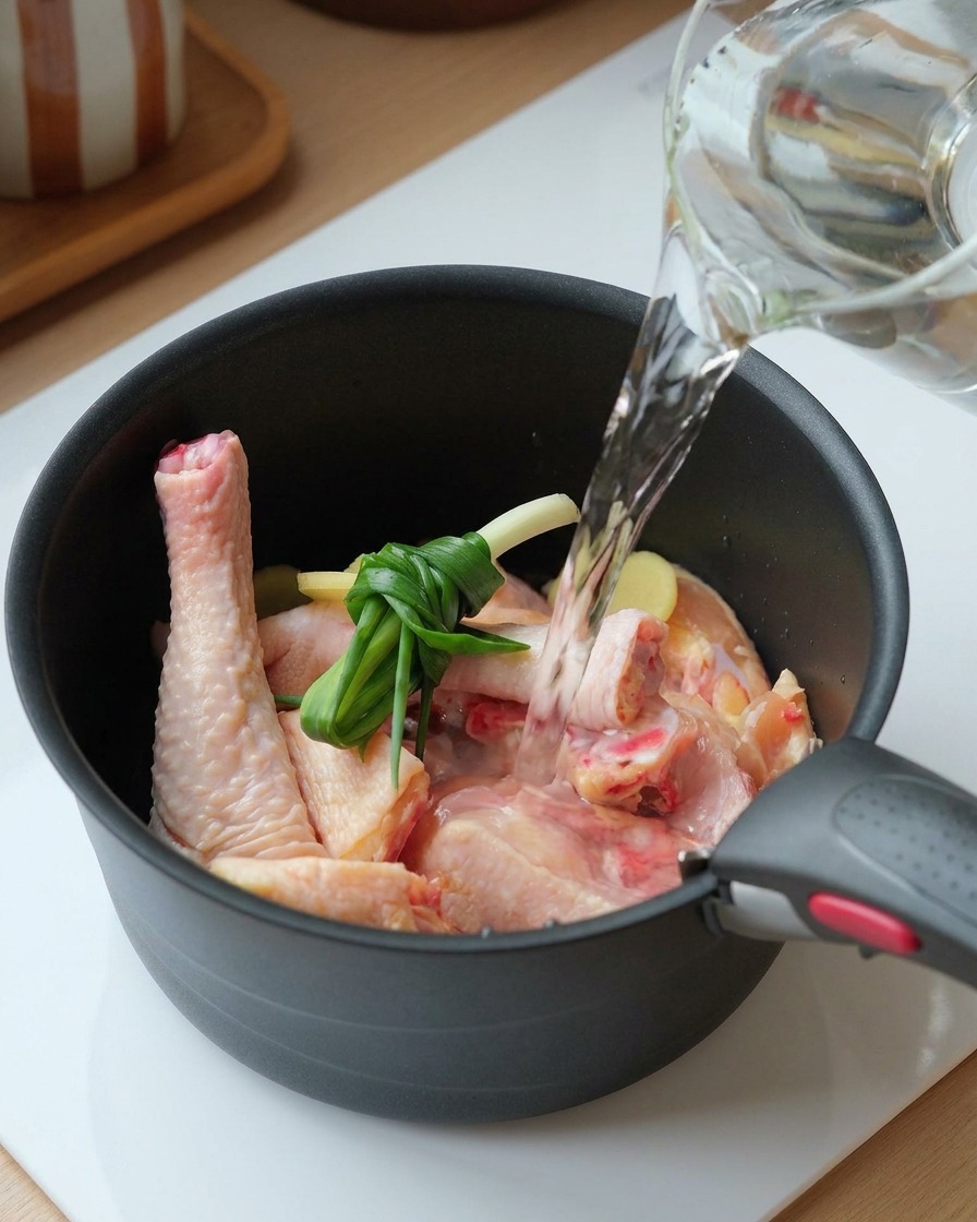 Acqua limpida versata da una caraffa di vetro in una pentola nera con pollo crudo, cipollotti legati e fette di zenzero.