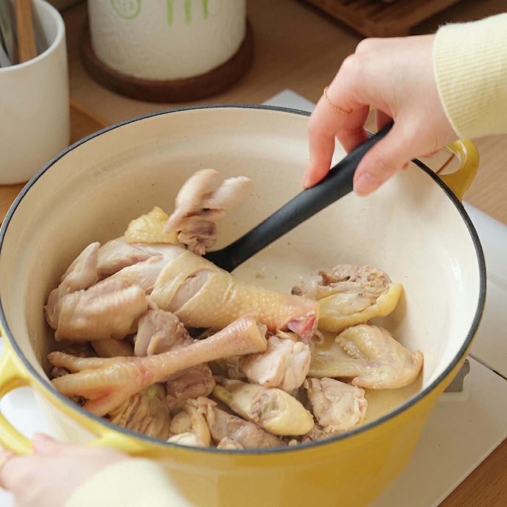 Una mano che usa una spatola nera per saltare i pezzi di pollo sbollentati in una pentola di ghisa gialla.