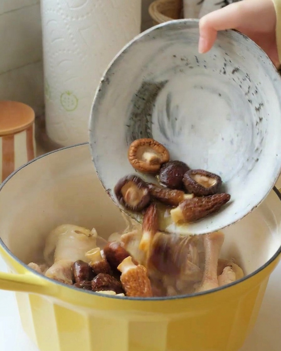Una ciotola di funghi shiitake e morelle ammollati versata in una pentola gialla con pezzi di pollo.