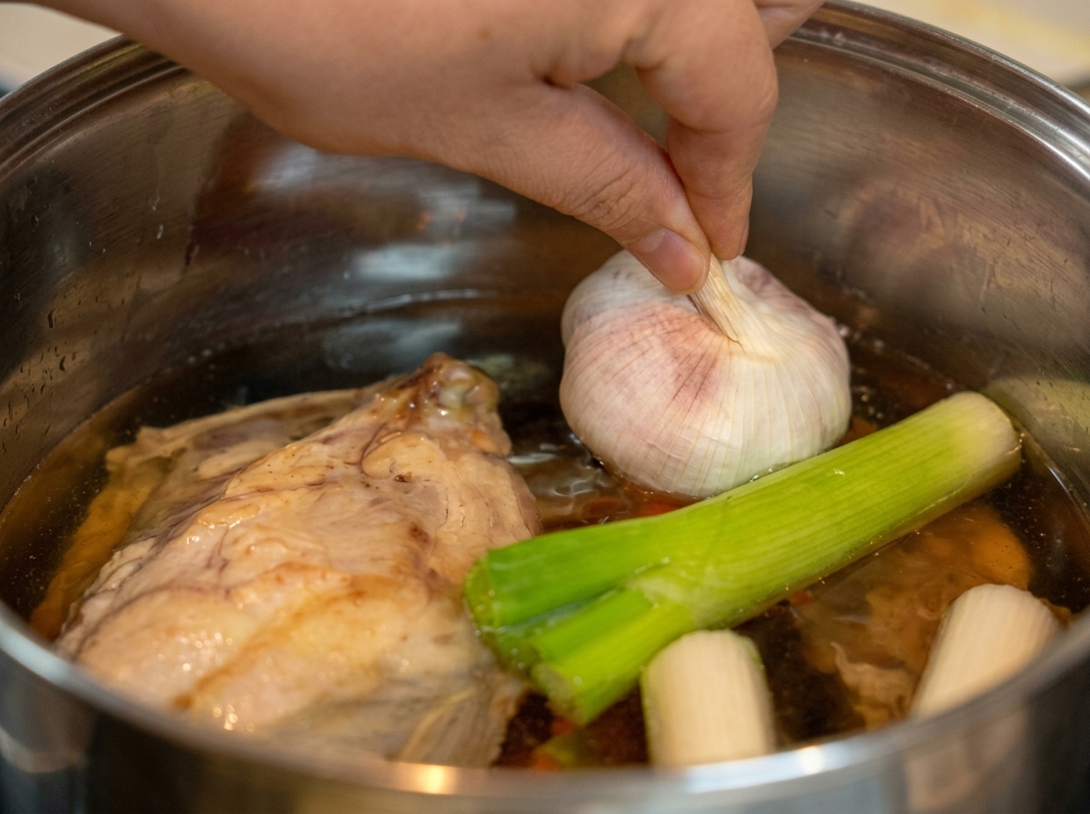 Una mano che mette una testa daglio intera in una pentola con acqua, pollo e porri.