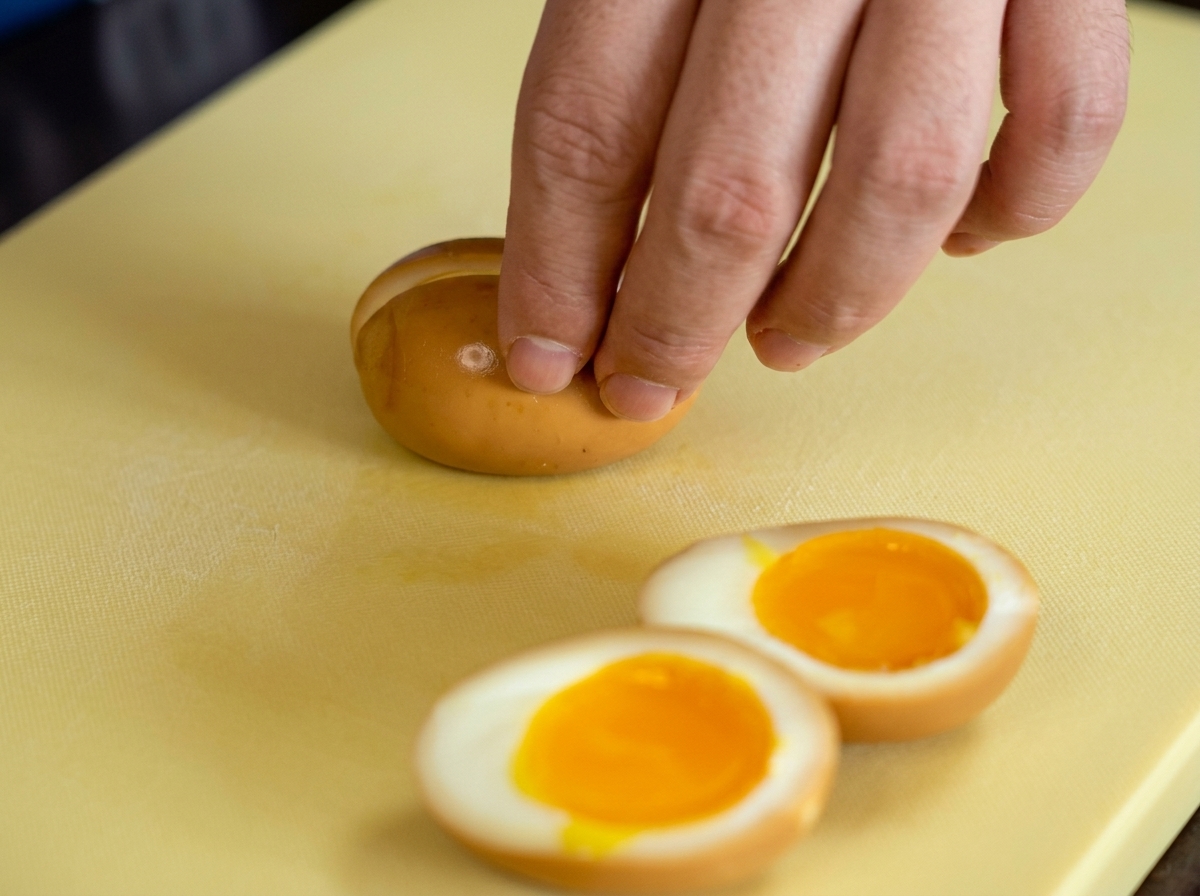 Una mano che taglia a metà un uovo marinato su un tagliere giallo, rivelando il tuorlo cremoso.