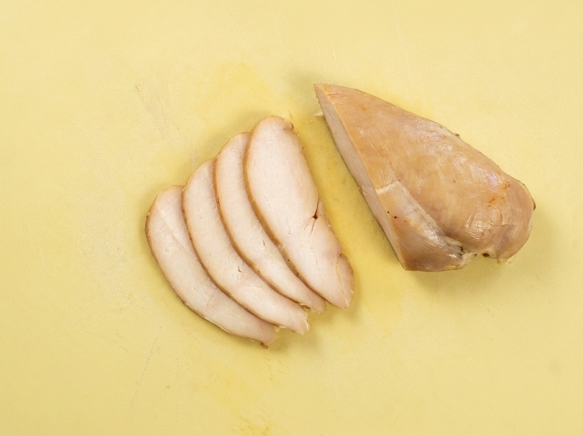 Fette sottili e uniformi di petto di pollo cotto disposte su un tagliere giallo.
