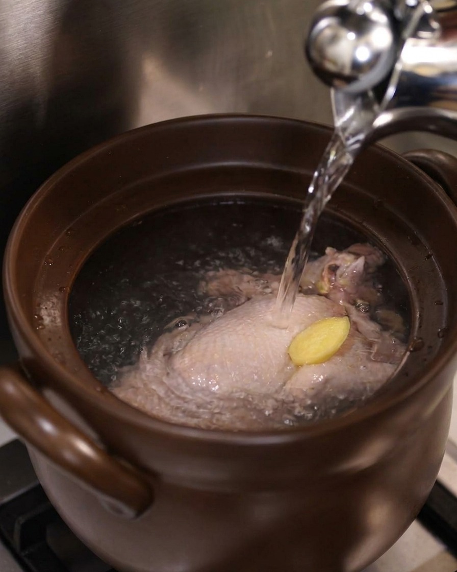 Acqua bollente versata da un bollitore dargento sopra un pollo intero e una fetta di zenzero in una pentola di ceramica.