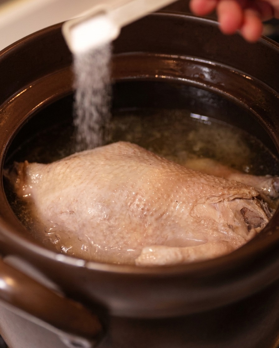 Una mano che usa un cucchiaino bianco per spargere sale sopra un pollo cotto nel brodo limpido.