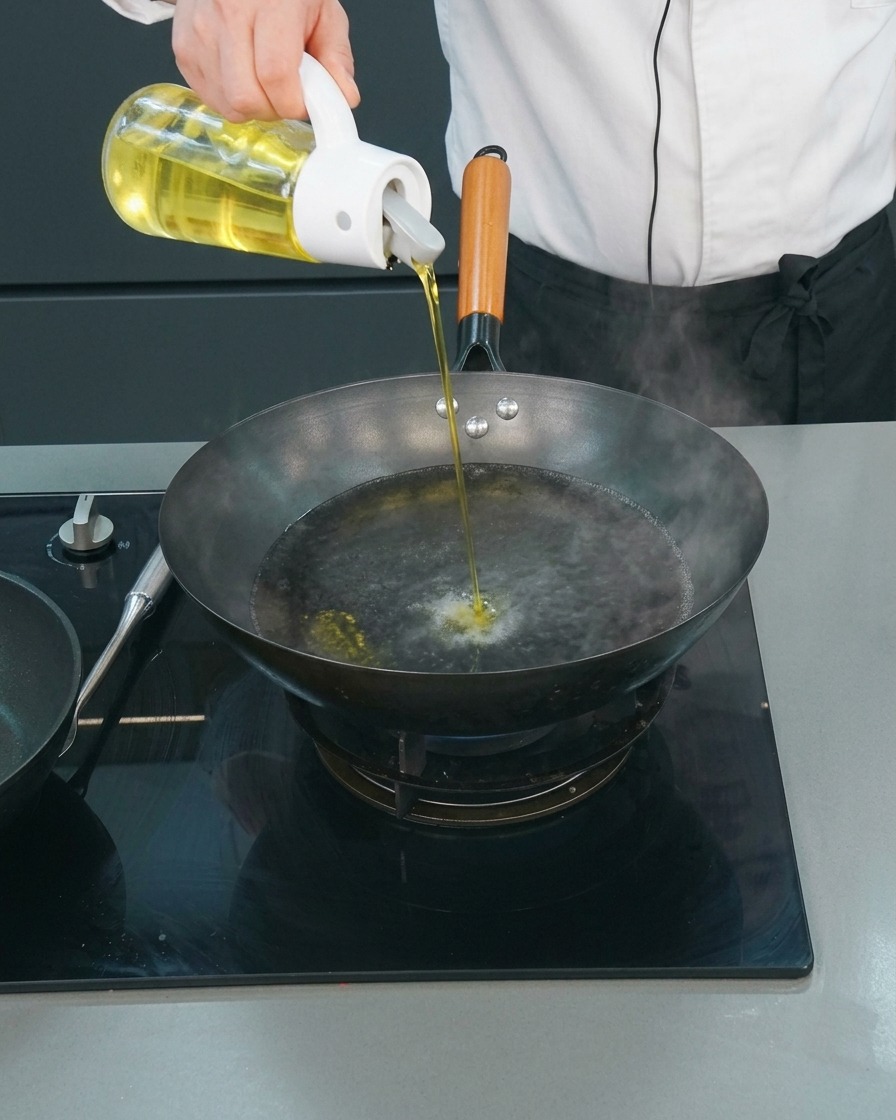 Uno chef che versa un filo dolio di semi da un dosatore di vetro in un wok con acqua bollente.