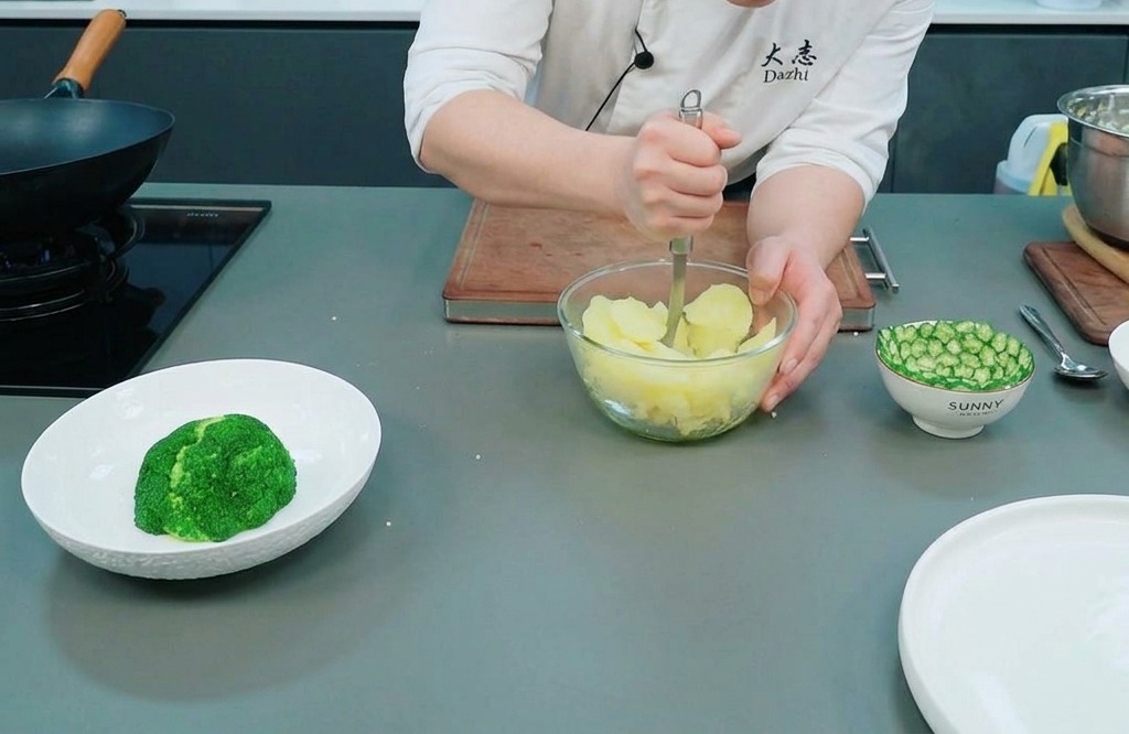 Chef che usa uno schiacciapatate in metallo per ridurre in purea le patate cotte al vapore in una ciotola di vetro.