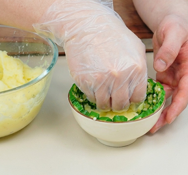 Una mano con guanto in plastica che preme il purè di patate in una ciotolina rivestita di fette di okra.