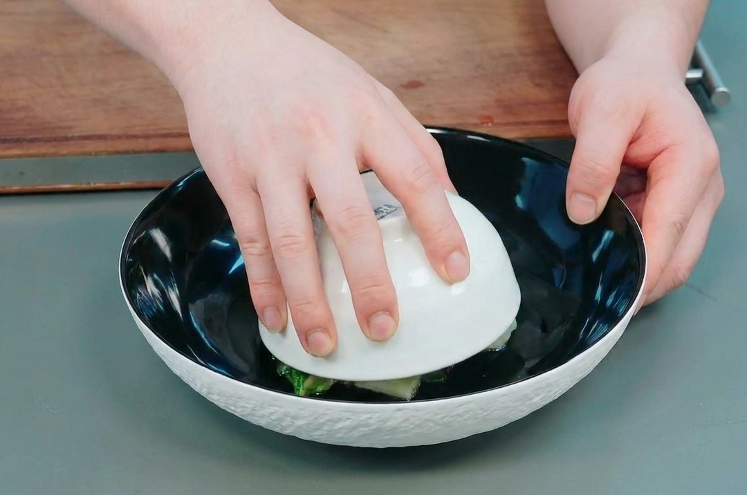 Mani che sollevano una ciotolina bianca da un piatto nero per rivelare la cupola di okra e patate.