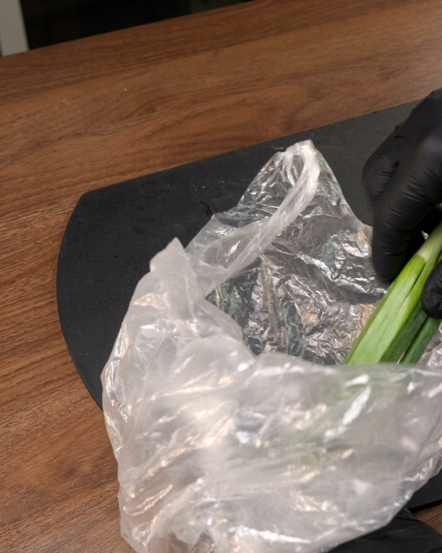 Una mano guantata che estrae verdura verde fresca da un sacchetto di plastica trasparente su una superficie scura.