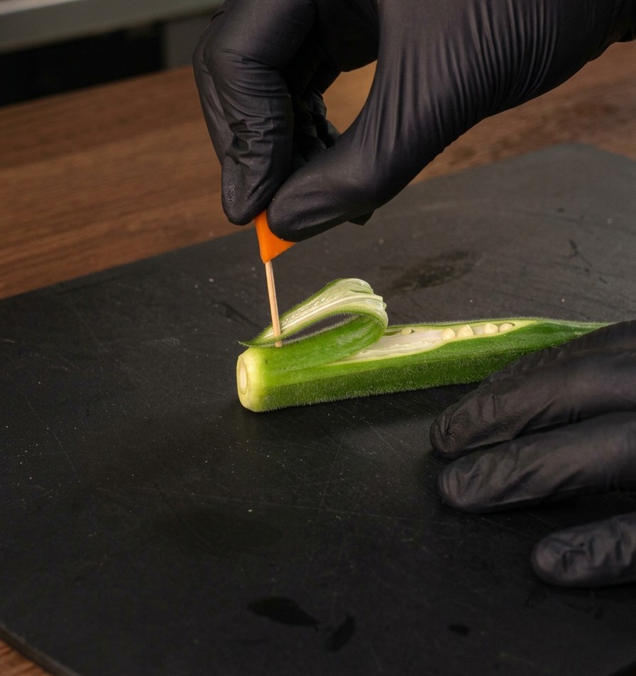Una mano guantata che inserisce una piccola bandiera arancione su uno stuzzicadenti nella vela di okra.