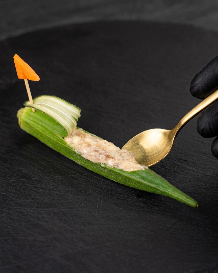 Un cucchiaino dorato che riempie un pezzo di okra svuotato con pasta di gamberi cruda, decorato con una bandierina di carota.