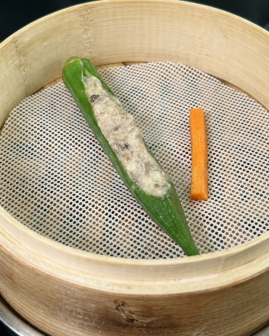 Un singolo baccello di okra farcito e un bastoncino di carota su una rete bianca allinterno di un cestello per il vapore in bambù.