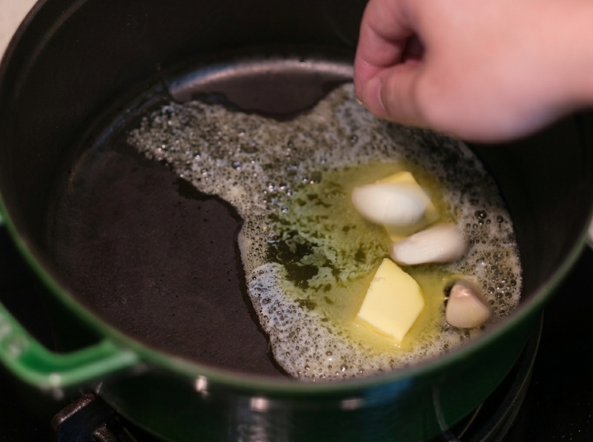 Una mano che mette spicchi daglio nel burro fuso spumeggiante dentro una pentola di ghisa verde.