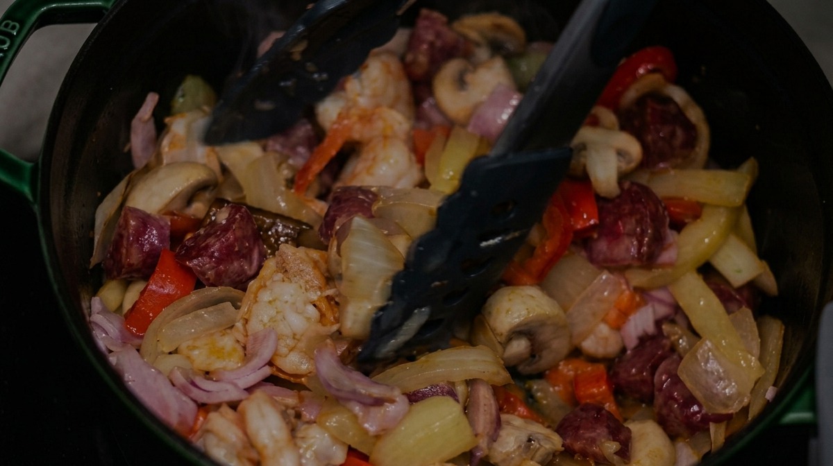 Mescolare salsiccia affumicata a fette, bacon e gamberi in una pentola di verdure miste con pinze nere.