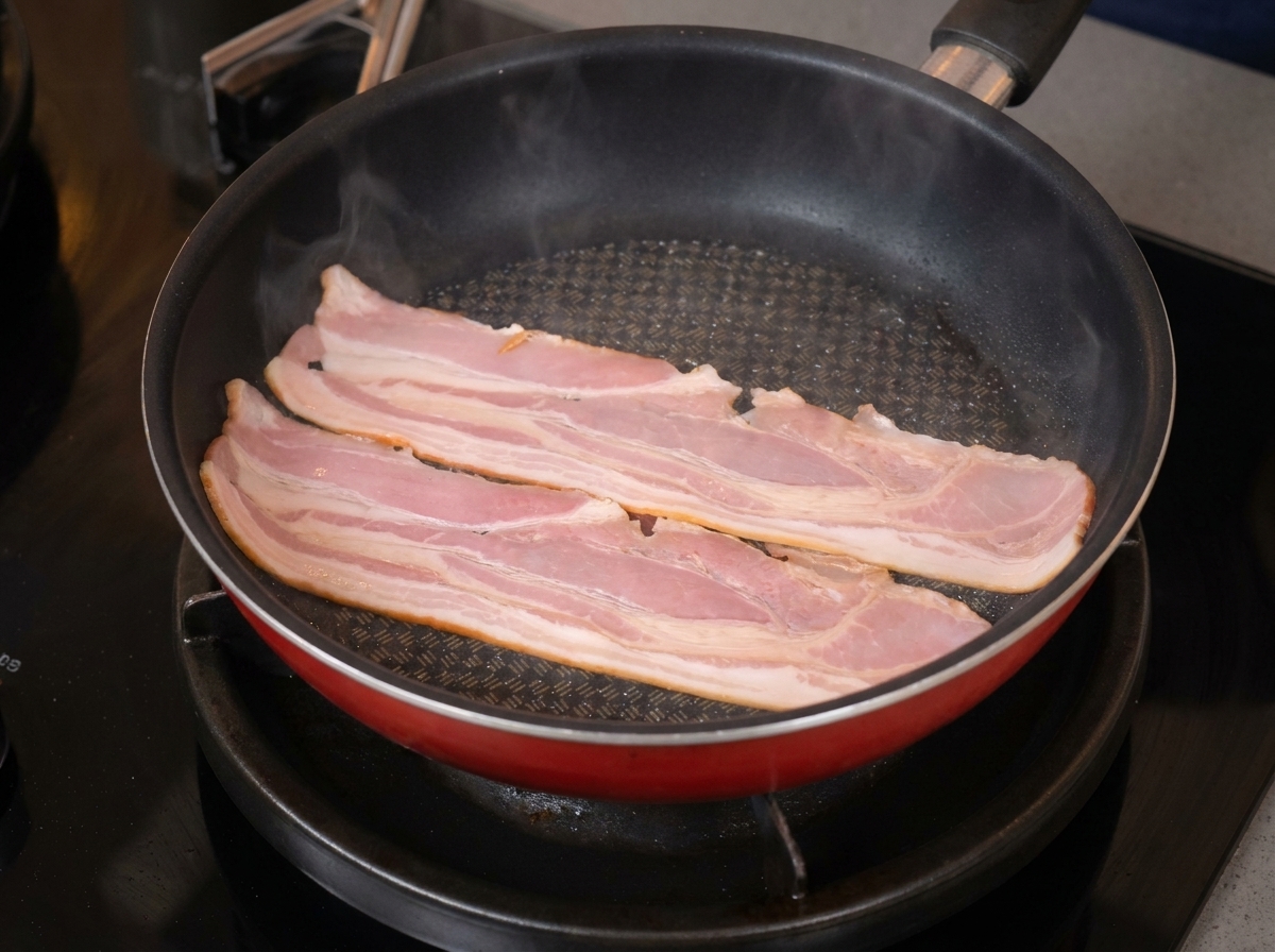 Due fette lunghe di pancetta che sfrigolano e iniziano ad arricciarsi in una padella antiaderente rossa.