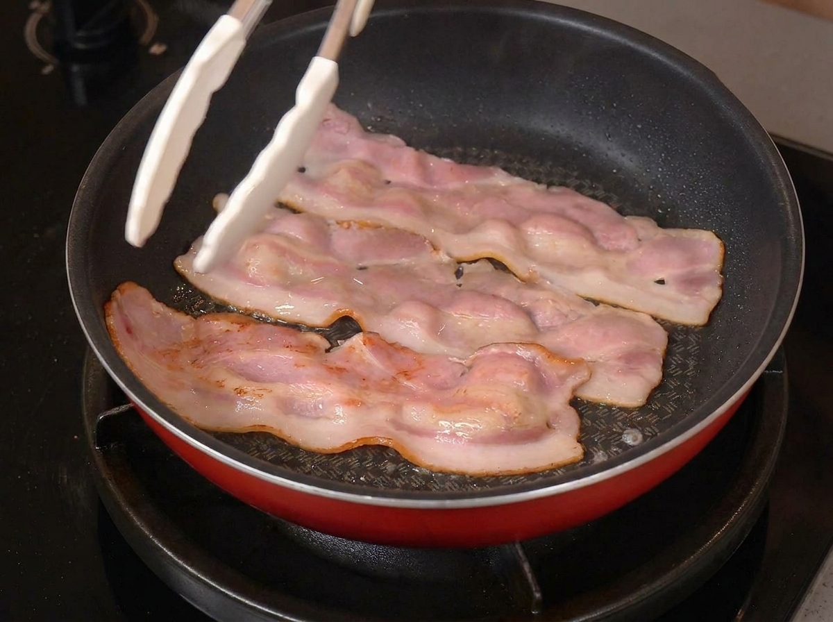 Tre strisce di pancetta che sfrigolano in padella con i bordi dorati, mentre una pinza bianca le afferra.